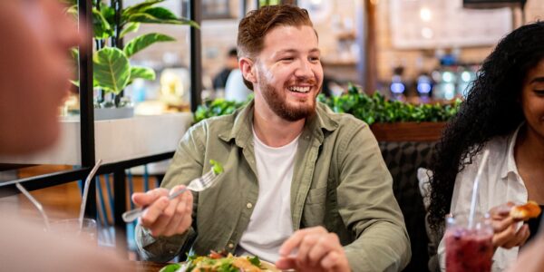 Feature image A young man at a dinner table in a restaurant. He is smiling and holding a fork over his healthy plate of food.